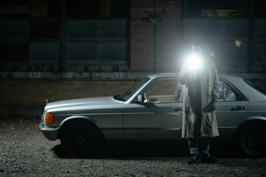 A detective stands by a vintage car at night, illuminated by a flashlight.