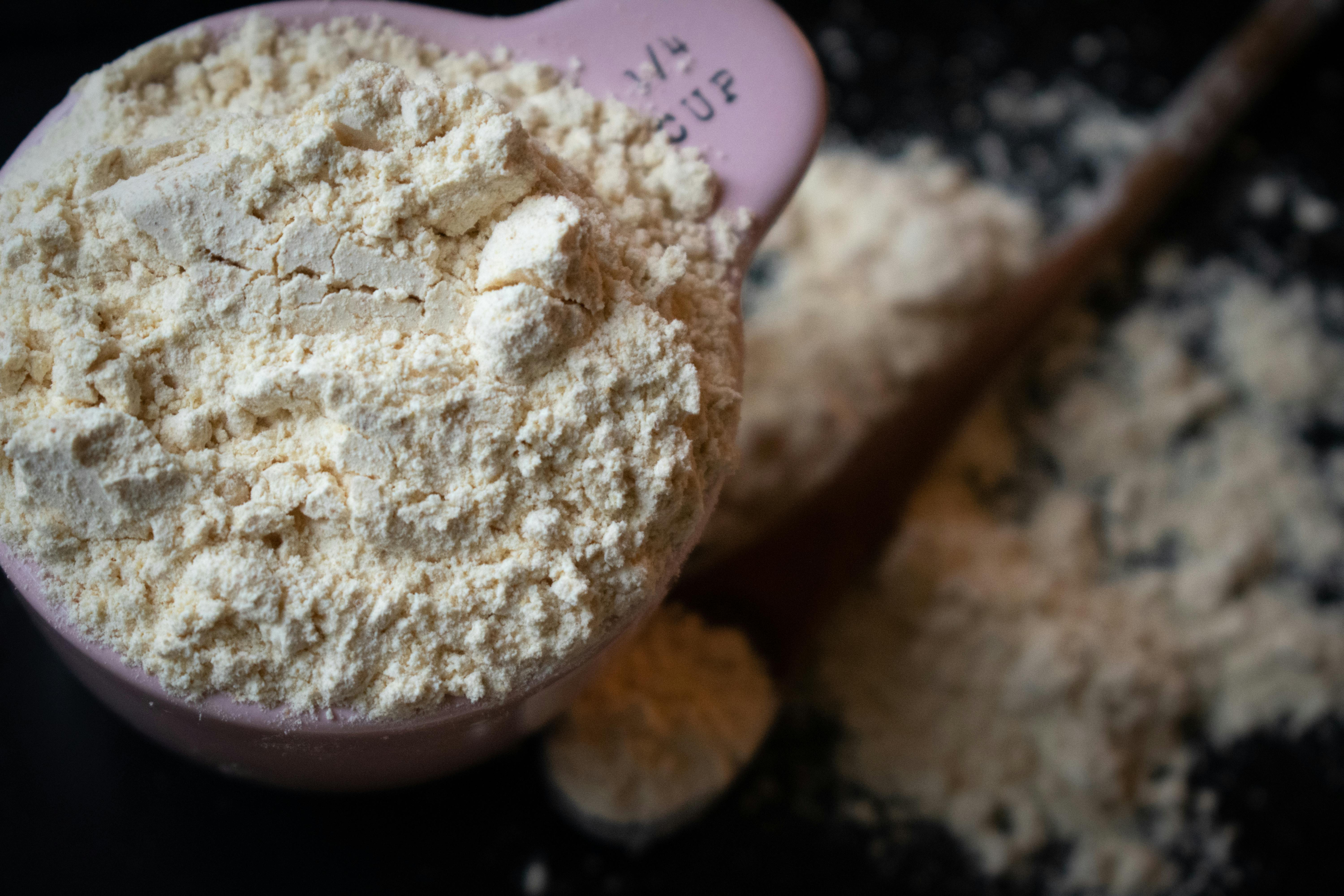 A filled measuring cup of flour spills onto a dark surface in a shallow depth of field.