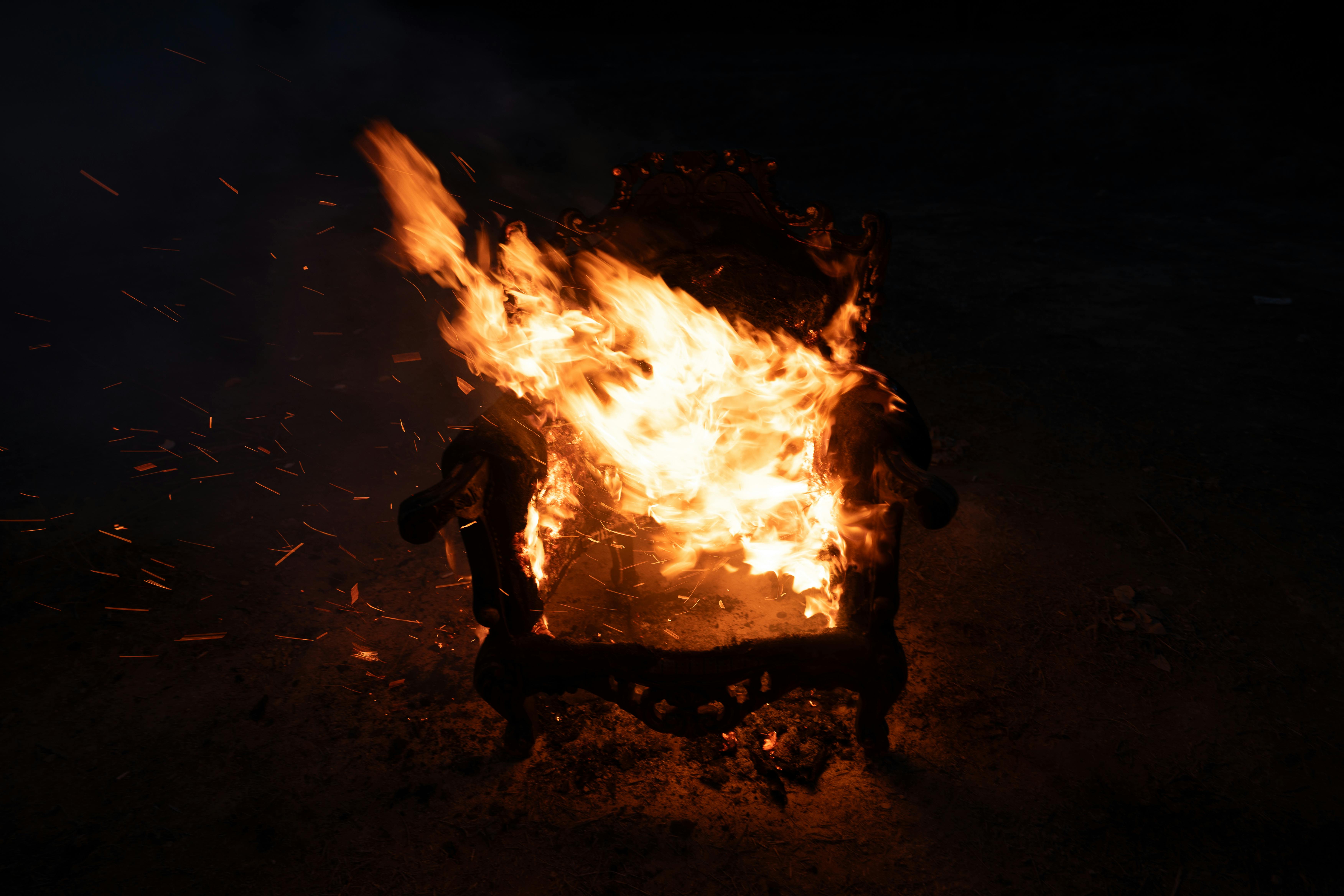 A dramatic scene of a wooden chair engulfed in flames at night, showcasing a vivid display of fire in a dark outdoor setting.