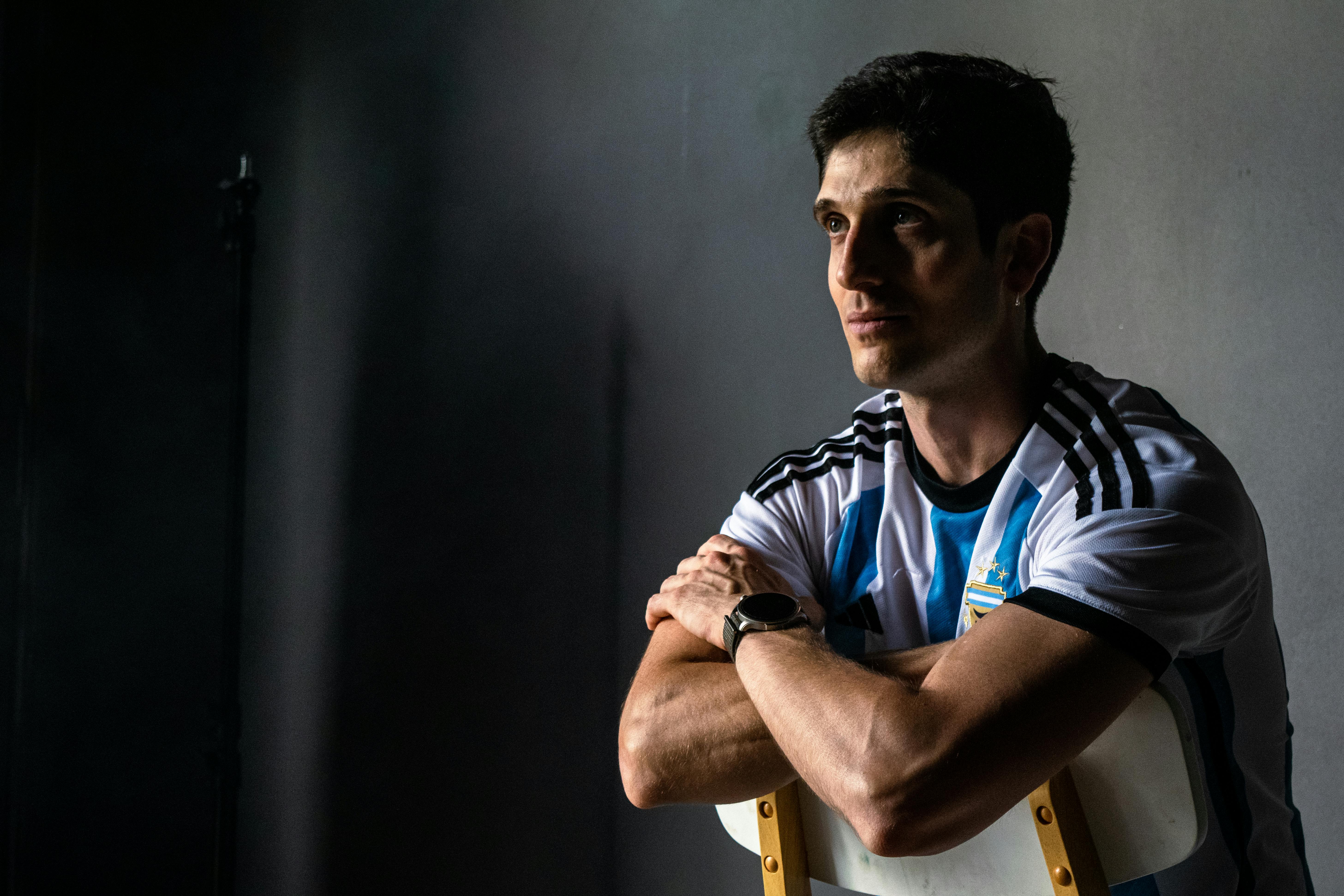 Reflective portrait of a man in an Argentina soccer jersey sitting indoors.