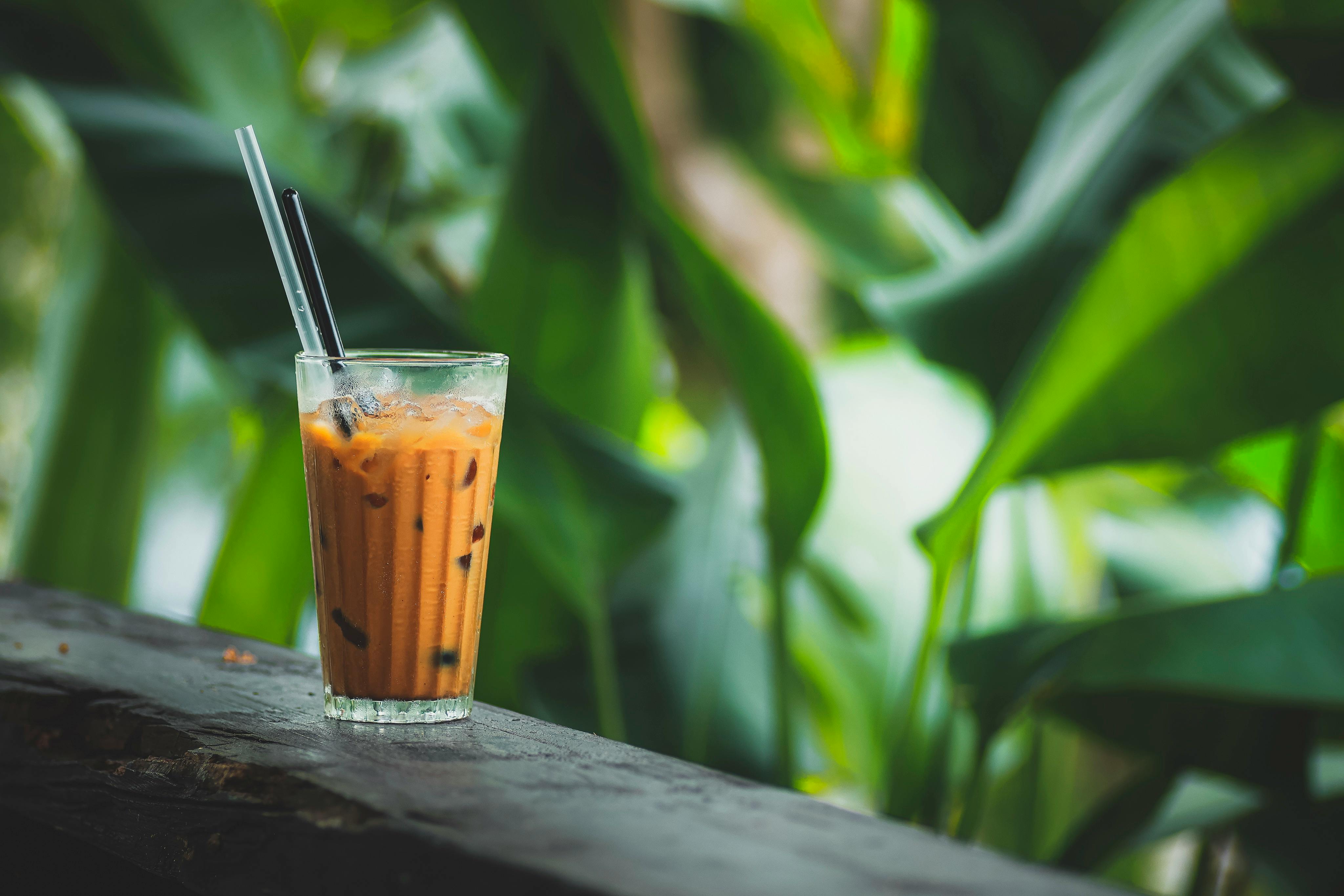 Glass of iced Thai tea with black straws against a lush green background. Perfect for summer vibes.